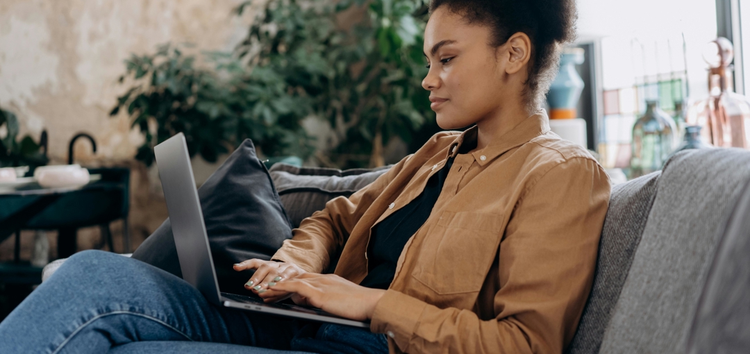 Een vrouw zit op een bank met een laptop op schoot, geconcentreerd aan het werk.